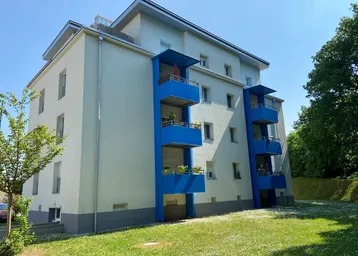 Modernes Mehrfamilienhaus mit hellen Fassaden und markanten blauen Balkonen, umgeben von einer gepflegten Grünfläche.