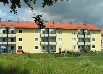 Mehrfamilienhaus mit gelber Fassade, rotem Ziegeldach und mehreren Balkonen, umgeben von einer grünen Wiese.