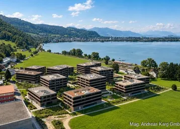 Luftaufnahme einer modernen Wohnanlage am Seeufer mit grünen Wiesen und Bergpanorama im Hintergrund.