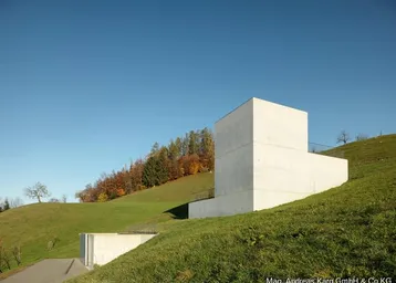 Modernes Gebäude aus Sichtbeton in einer grünen Hügellandschaft unter blauem Himmel.
