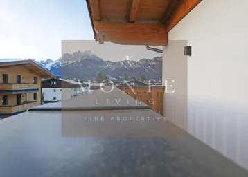 Blick von einem Balkon mit Holzgeländer auf schneebedeckte Berge und umliegende Häuser in alpiner Umgebung.