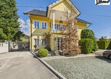Gelbes Einfamilienhaus mit Säulen am Eingang und gepflegtem Vorgarten unter blauem Himmel.
