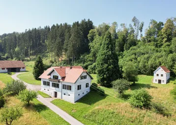 Ländliches Anwesen mit mehreren Gebäuden, umgeben von grünen Wiesen und Wäldern unter blauem Himmel.