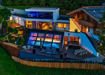 Luxuriöses Anwesen bei Nacht mit beleuchtetem Pool, großer Terrasse und Spielplatz in alpiner Umgebung.