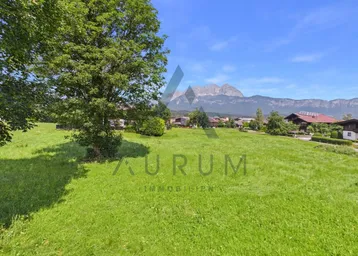 Weitläufige grüne Wiese mit Bergblick und traditionellen Häusern im Hintergrund unter blauem Himmel.