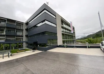 Modernes Gebäude mit dunkler Fassade, Balkonen und Ladestationen für Elektrofahrzeuge im Vordergrund.