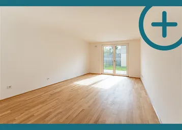 Helles, leeres Zimmer mit Parkettboden und Zugang zu einem kleinen Gartenbereich.