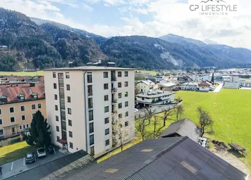 Mehrstöckiges Wohngebäude in ländlicher Umgebung mit Bergen im Hintergrund und Parkplätzen davor.