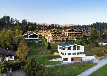 Panoramablick auf eine Ansammlung von modernen und traditionellen Häusern in einer bewaldeten Hanglage mit Bergblick.