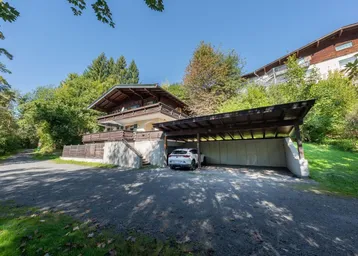 Haus mit Carport und Zufahrt, umgeben von Bäumen an einem sonnigen Tag.