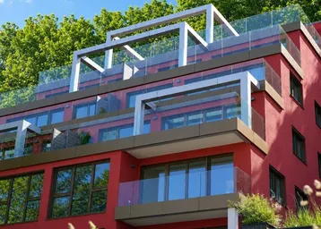 Modernes Wohngebäude mit roter Fassade, Glasbalkonen und grüner Umgebung unter blauem Himmel.