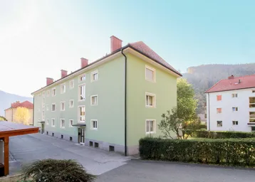 Grünes Mehrfamilienhaus mit rotem Ziegeldach und umliegender Hecke in bergiger Landschaft.