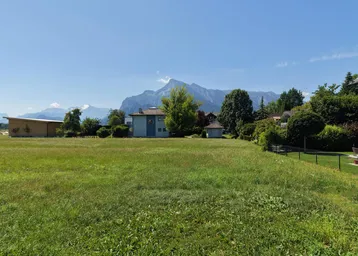 Weitläufige Grünfläche mit Blick auf die umliegende Berglandschaft und mehrere Wohnhäuser unter blauem Himmel.