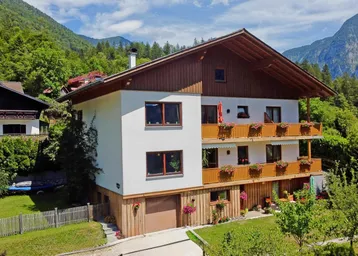 Traditionelles Haus im alpinen Stil mit Holzbalkonen und Bergblick, umgeben von grüner Natur.