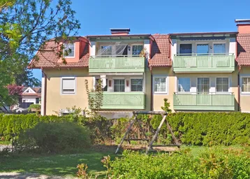 Mehrfamilienhaus mit mehreren Balkonen, Ziegeldach und einem kleinen Spielplatz im Vorgarten.