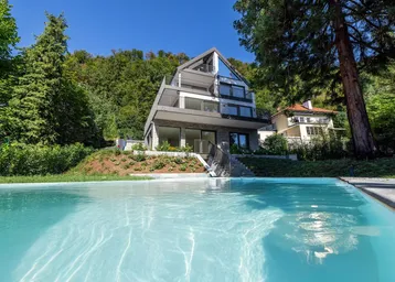 Modernes Haus mit Pool und Bergblick, umgeben von viel Grün und Bäumen.