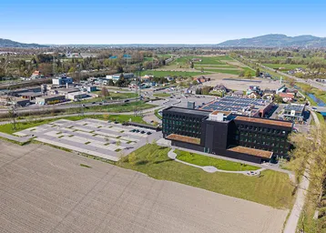 Luftaufnahme eines modernen Bürogebäudes mit umliegenden Feldern und Parkplätzen unter blauem Himmel.