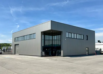 Modernes Geschäftsgebäude mit grauer Fassade und großen Glasflächen, umgeben von einem Parkplatz unter blauem Himmel.