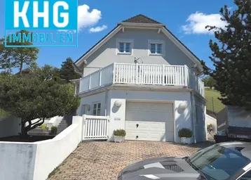 Graues Einfamilienhaus mit Garage und Balkon, umgeben von Grünflächen unter blauem Himmel.