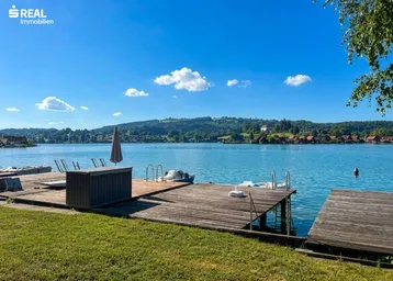 Idyllischer Blick auf den See mit privatem Holzsteg und Liegeflächen unter blauem Himmel.