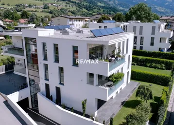 Moderne Wohnanlage mit weißer Fassade, Balkonen, Solarpaneelen und Bergblick in grüner Umgebung.