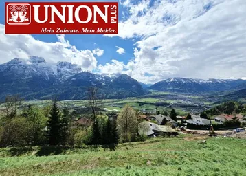 Panoramablick auf schneebedeckte Berge und eine grüne Landschaft mit Wohnbebauung unter blauem Himmel.