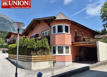 Hausfassade in lachsfarbenem Putz mit Erker und Holzbalkonen vor alpiner Bergkulisse.