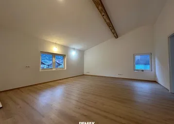 Geräumiger, leerer Raum mit Holzboden, Dachschräge und Fenstern mit Blick auf die Umgebung.