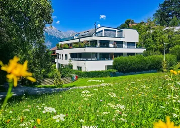 Modernes Mehrfamilienhaus mit Terrassen und Bergblick, umgeben von grüner Wiese und Bäumen.