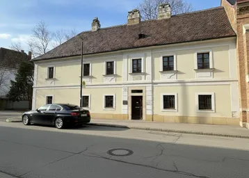 Historisches Gebäude mit beiger Fassade, braunen Fenstern und Satteldach an einer ruhigen Straße.