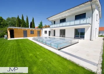 Modernes Haus mit großem Garten, überdachtem Pool und Holz-Nebengebäude unter blauem Himmel.