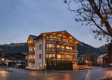 Modernes Mehrfamilienhaus im alpinen Stil mit Holzfassade und Balkonen in abendlicher Beleuchtung.