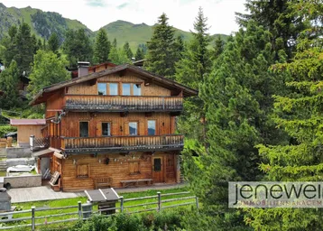 Traditionelles Holzhaus in alpiner Umgebung, umgeben von grünen Bäumen und Bergen.
