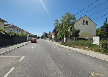 Blick auf eine ruhige Wohnstraße mit Häusern und einem sonnigen Baugrundstück unter blauem Himmel.
