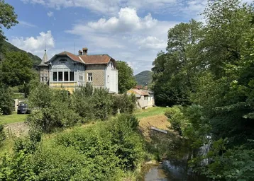 Historische Villa am Flussufer, umgeben von üppigem Grün und Bergen im Hintergrund.