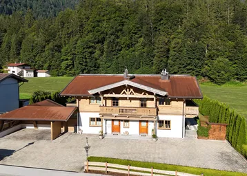 Traditionelles Haus im alpenländischen Stil mit Carport und gepflastertem Vorplatz, umgeben von grünen Wiesen und Wäldern.