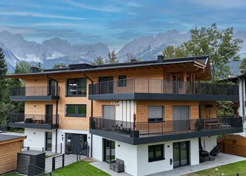 Modernes Mehrfamilienhaus mit Holzfassade und mehreren Balkonen vor alpiner Bergkulisse.