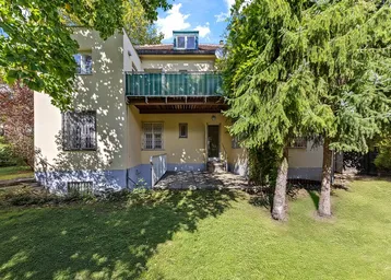 Hinteransicht eines gepflegten Hauses mit grünem Garten, gepflasterter Terrasse und einem Balkon.