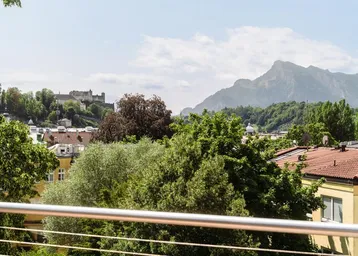 Blick von der Terrasse über die Stadt Salzburg mit Festung Hohensalzburg und umliegenden Bergen.
