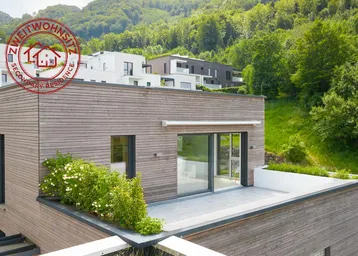 Moderne Architektur mit Holzfassade und großer Dachterrasse, umgeben von grünen Hügeln.