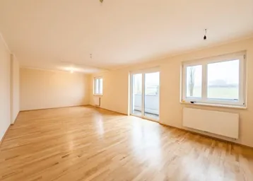Heller, leerer Raum mit Holzfußboden, großen Fenstern und Zugang zu einem Balkon mit grüner Aussicht.
