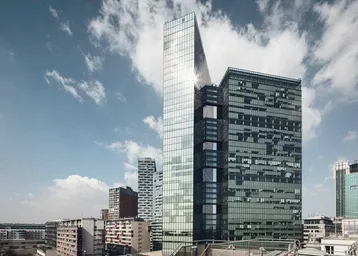 Moderne Glasfassade eines Hochhauses unter blauem Himmel mit Wolken, umgeben von städtischer Architektur.