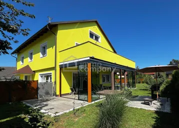 Gelbes Haus mit überdachter Terrasse, Grillbereich und gepflegtem Garten unter blauem Himmel.