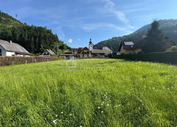 Weitläufige grüne Wiese mit Blick auf ein Dorf und die umliegenden Berge unter blauem Himmel.