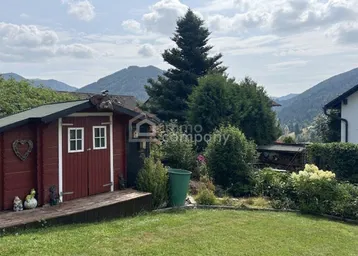 Roter Gartenschuppen in einem gepflegten Garten mit Rasen und hohen Bäumen im Hintergrund.