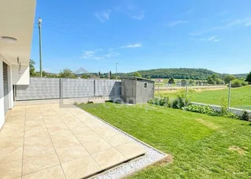 Moderne Terrasse mit Fliesenboden und angrenzendem Rasen, umgeben von grüner Landschaft unter blauem Himmel.