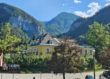 Gelbes Gebäude mit Mansarddach, umgeben von grünen Bäumen und majestätischen Bergen unter blauem Himmel.