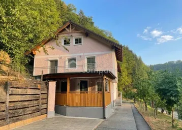Hausfassade mit Balkonen und Holzelementen, eingebettet in eine grüne Hügellandschaft unter blauem Himmel.