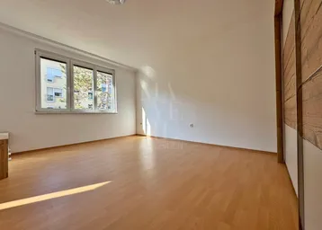 Helles, leeres Zimmer mit Parkettboden und großem Fenster, das viel Tageslicht hereinlässt.