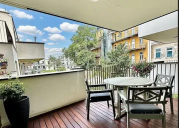 Geräumiger Balkon mit Holzboden und Sitzgelegenheiten, umgeben von städtischem Grün und Gebäuden.